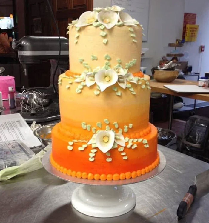 Three-tiered orange gradient cake decorated with white icing flowers, displayed in a bakery kitchen setting.