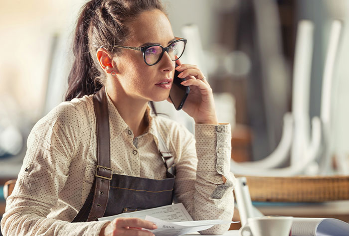 Woman bakery worker in apron and glasses talking on phone, holding papers, planning her revenge on bakery manager.
