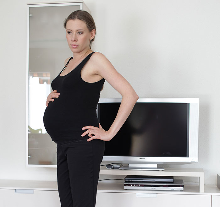 Pregnant woman standing with hand on belly and hip, looking concerned in a modern living room with TV behind her.