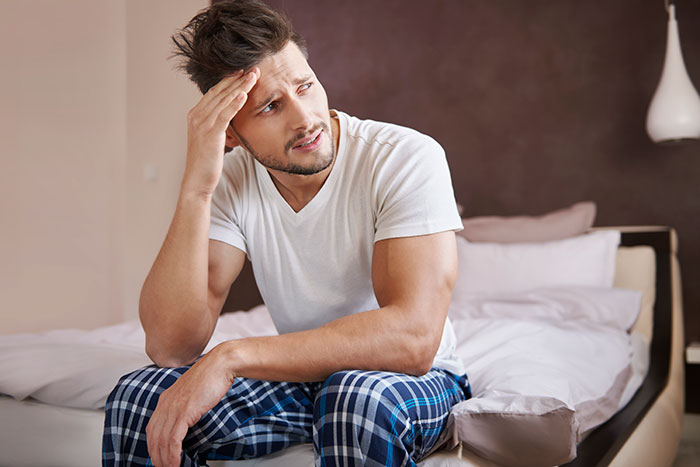 Man sitting on bed looking stressed and frustrated, wearing a white t-shirt and blue plaid pants in bedroom.