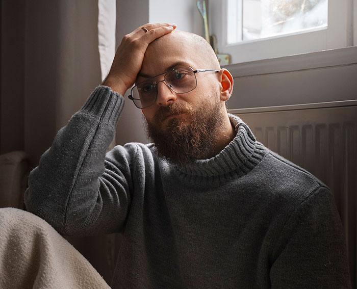 Man sitting indoors looking frustrated and stressed, wearing glasses and a gray sweater near a window.
