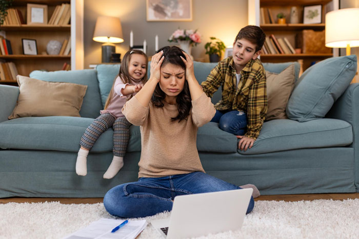 Stressed mom of 3 sitting on floor with laptop while children play on couch behind her in cozy living room.