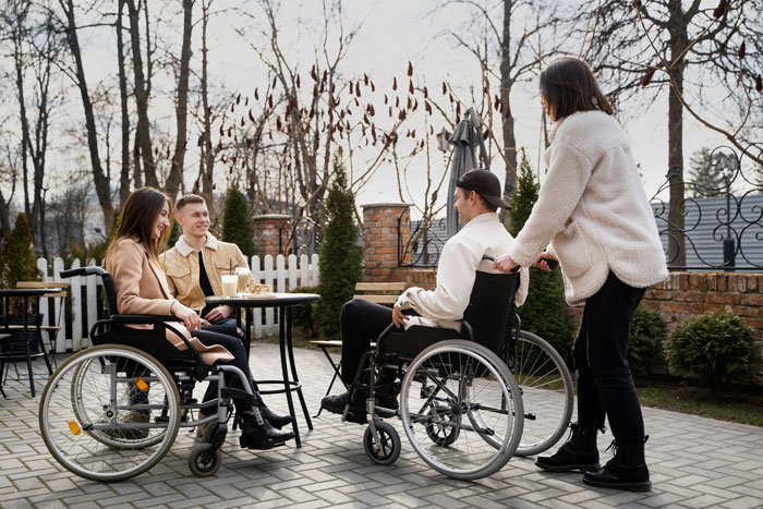 Mom of 3 sitting outdoors in a wheelchair while a woman pushes another person in a wheelchair in a social setting.