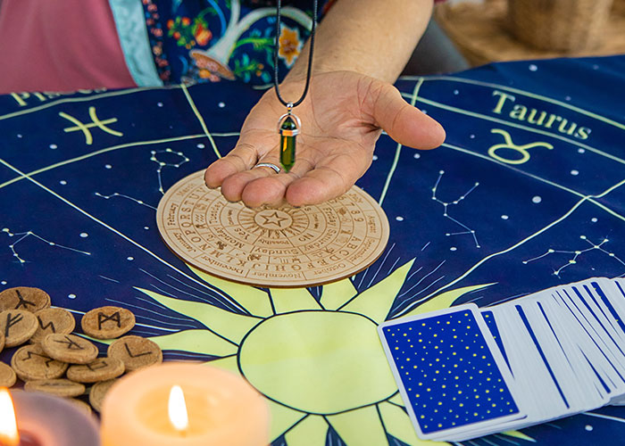 Person holding a pendulum over zodiac chart with tarot cards and runes, illustrating psychic predictions coming true.