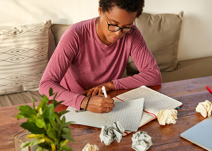 Young person wearing glasses writing notes at a wooden table surrounded by crumpled paper and notebooks about psychic predictions.