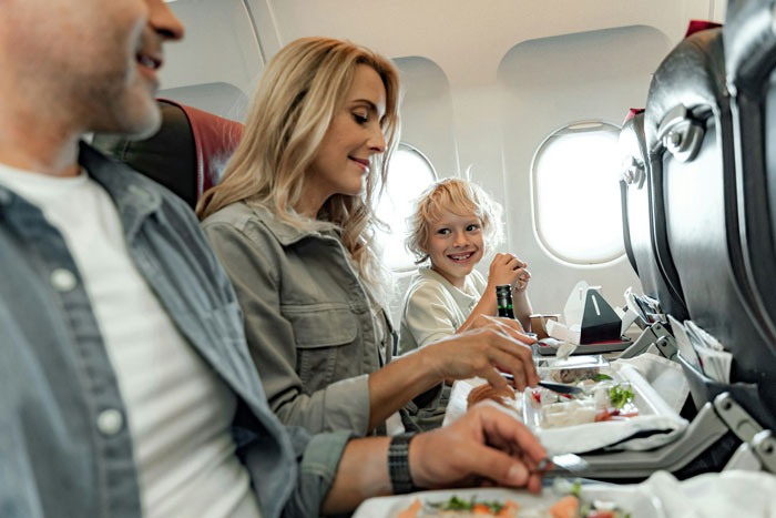 Family eating airplane meal while child looks at woman refusing to swap plane seat with rude family member