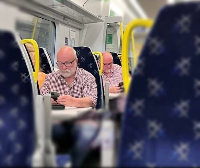 Man with glasses on train engrossed in phone, reflection in mirror creating an out of context humorous scene.