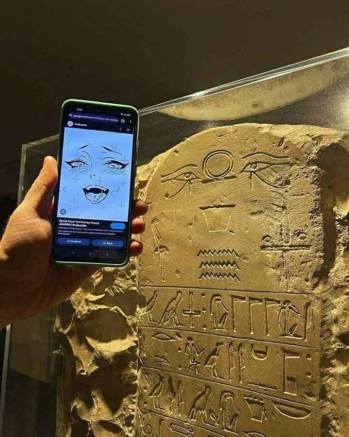 Hand holding phone showing anime facial expression next to ancient stone with occult symbols and hieroglyphics.