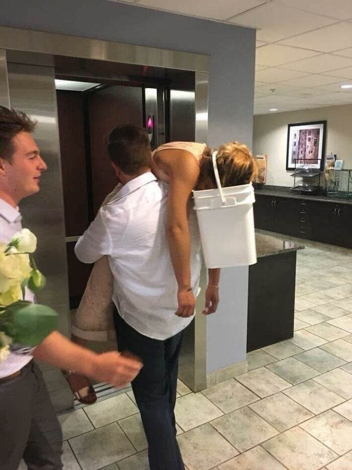 Man carrying unconscious woman with a bucket on her head, humorously caught out of context in an indoor setting.