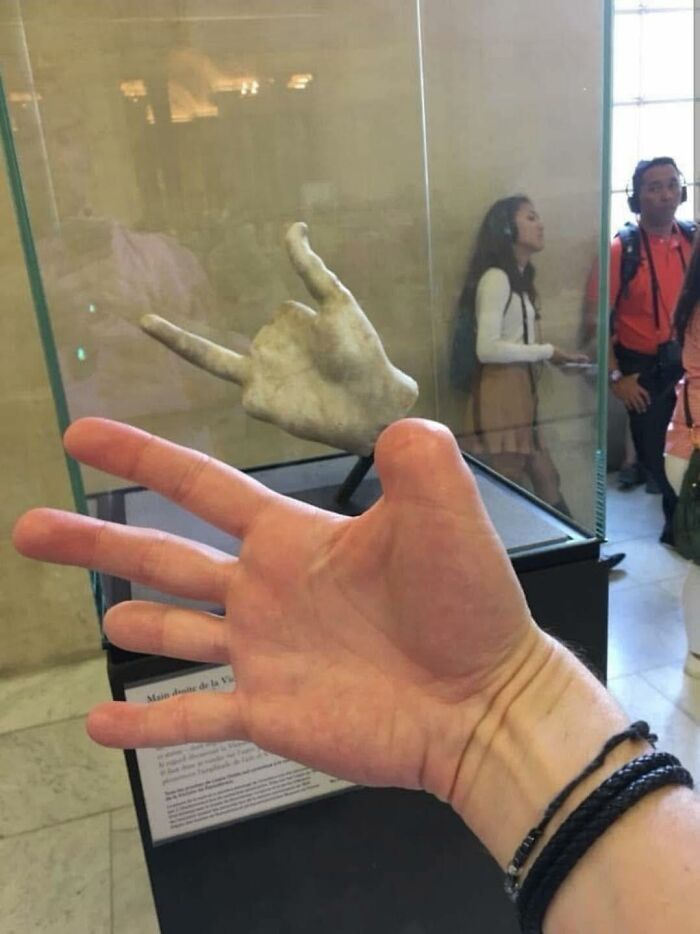 Stone hand sculpture making a horned gesture behind a real hand in a museum, illustrating pleasures of the occult humor.