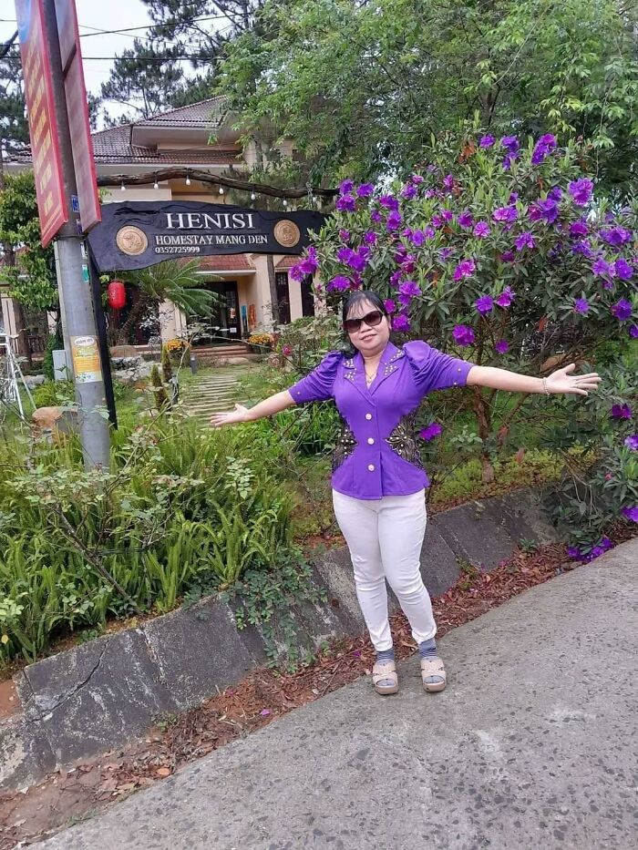 Woman in a purple top posing with arms outstretched near purple flowers outside a homestay, highlighting pleasures of the occult theme.