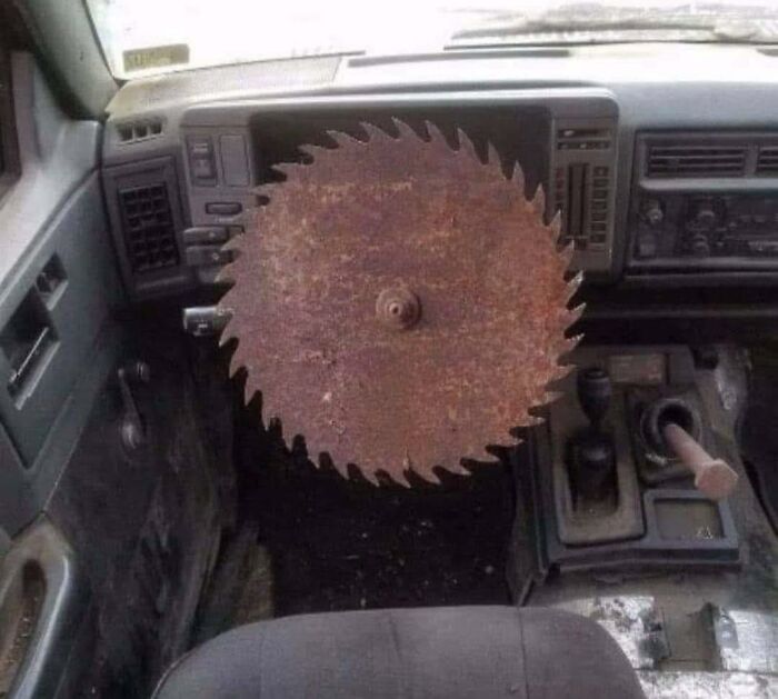 Rusty circular saw blade mounted inside a vehicle steering wheel area, creating a bizarre occult-themed scene.