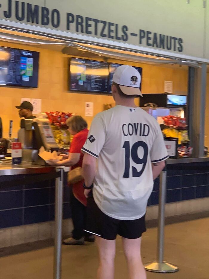 Person wearing a COVID 19 jersey standing in line at a concession stand for jumbo pretzels and peanuts, capturing pleasures of the occult humor.