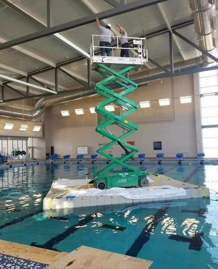 Two workers on an elevated platform over a floating base in a swimming pool, showing out of context pleasures of the occult humor.
