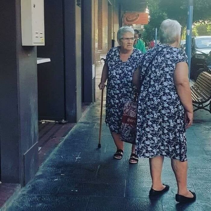 Two elderly women wearing identical floral dresses standing and walking on a city sidewalk, creating a humorous occult illusion.