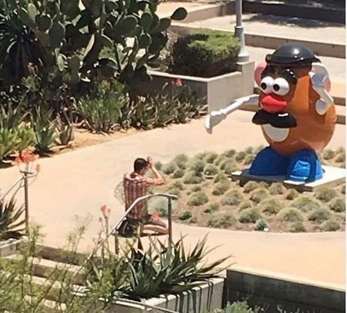 Man sitting outdoors next to a giant Mr. Potato Head statue, a humorous scene for pleasures of the occult pics.