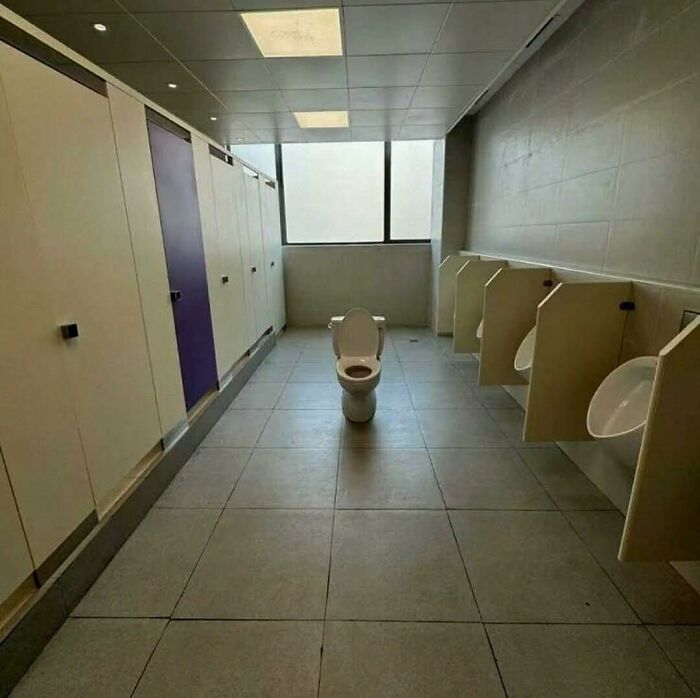 A lone toilet in the middle of a public restroom with stalls on one side and urinals on the other, out of context humor.