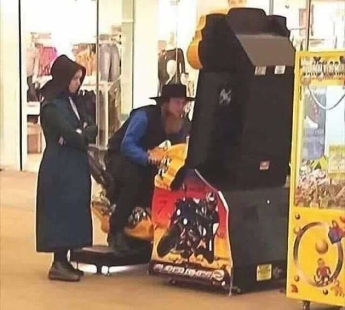 Two people in traditional occult clothing playing an arcade racing game in a public indoor space.