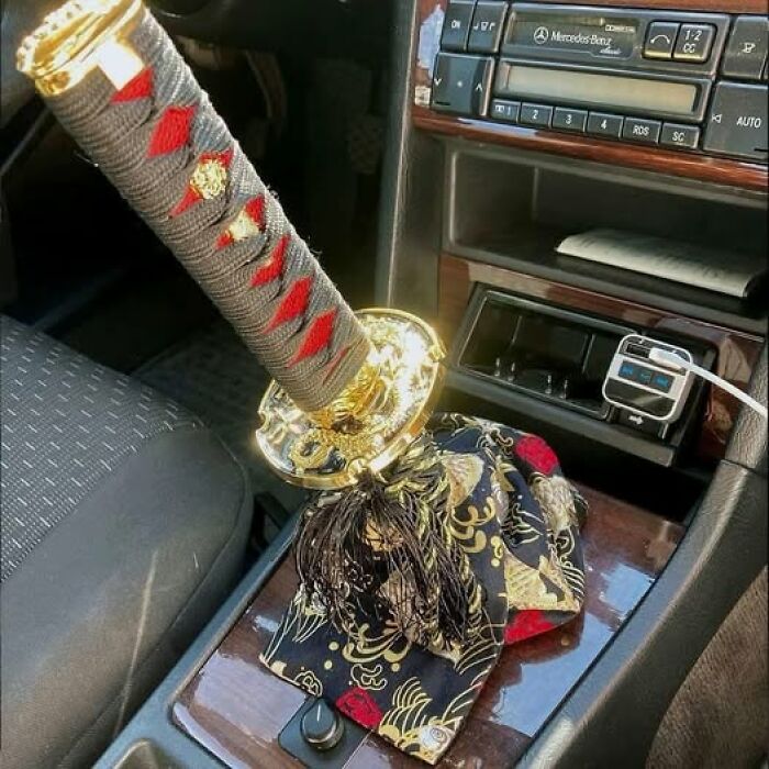 Katana sword gear shift k**b with intricate red and gold designs inside a car, blending pleasures of the occult with humor.
