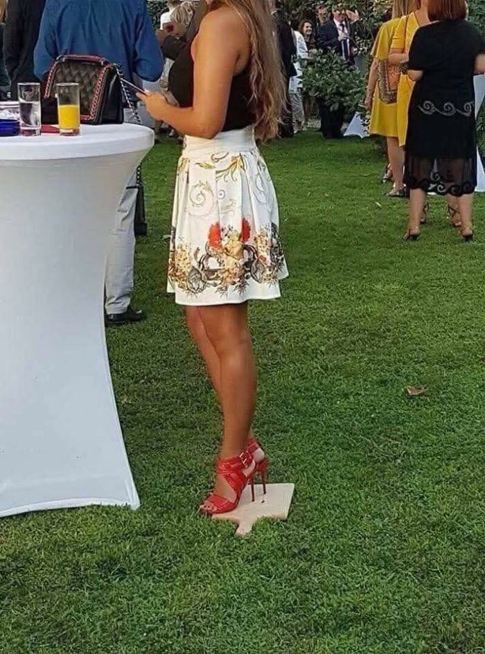 Woman wearing red heels standing on a wooden hand-shaped platform on grass at an outdoor event with people around.