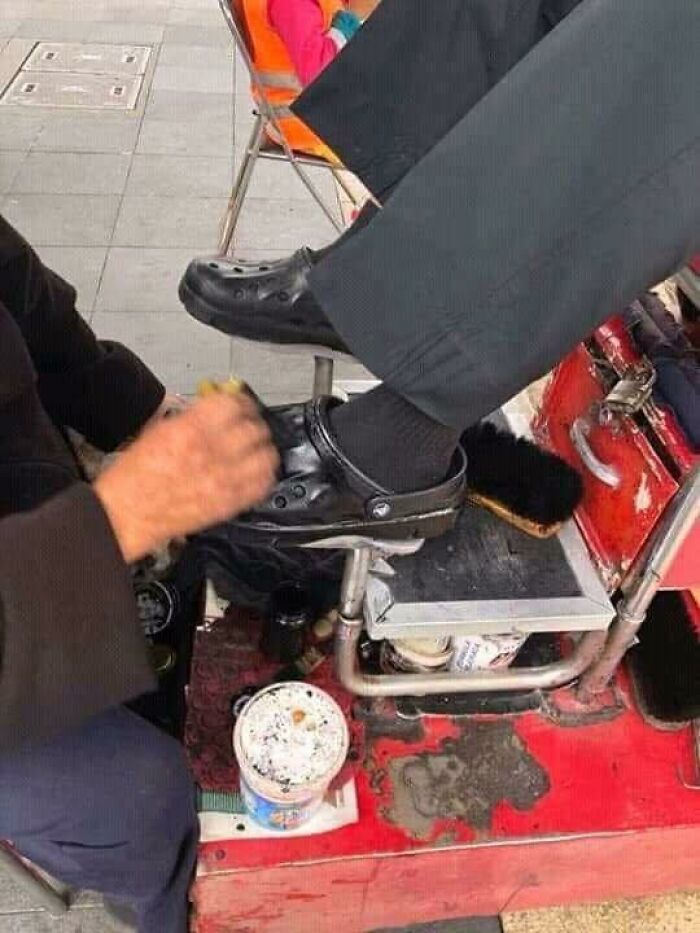 Shoe shiner polishing a pair of black Crocs on a street setup, highlighting pleasures of the occult humor in daily life.