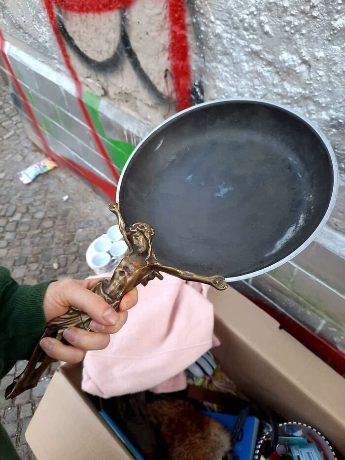 Hand holding a frying pan with a crucifix figure handle, combining occult imagery and an everyday object outdoors.