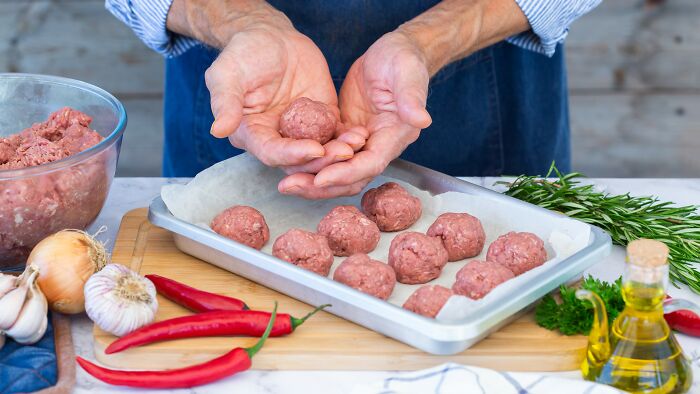 Man shaping raw meatballs by hand on a tray, surrounded by fresh ingredients including garlic, chili, and herbs.