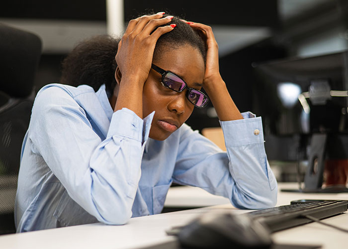 Stressed employee in glasses holding head at desk, reflecting on exposing management and HR to coworkers.