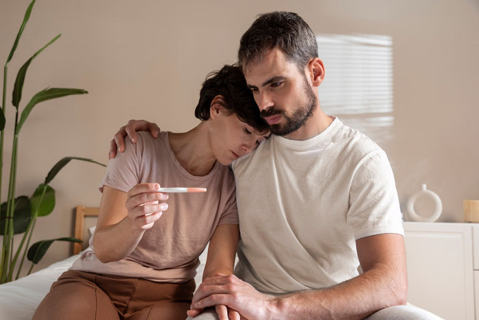 Sad couple sitting on bed with pregnancy test, struggling with infertility and family issues involving bio mom and stepmom. Sad couple sitting on bed with pregnancy test, struggling with infertility and family issues involving bio mom and stepmom.