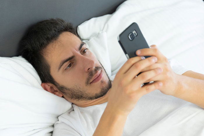 Man lying in bed looking serious while using smartphone, reflecting on refusing visiting girlfriend hospital.