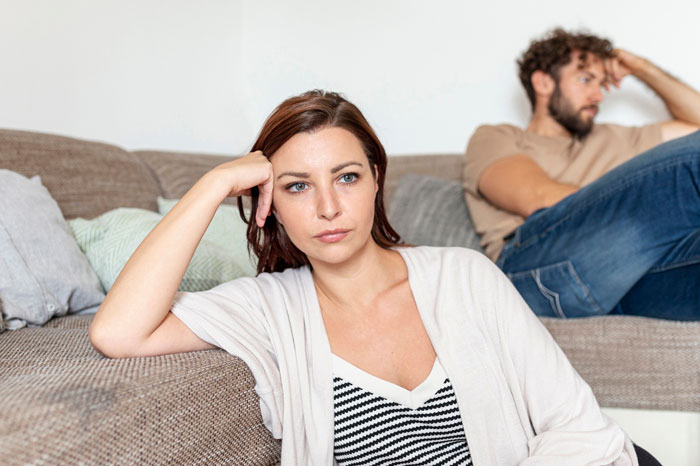 Frustrated wife sitting on couch with upset manchild playing games in background, tense family moment over lost dog.