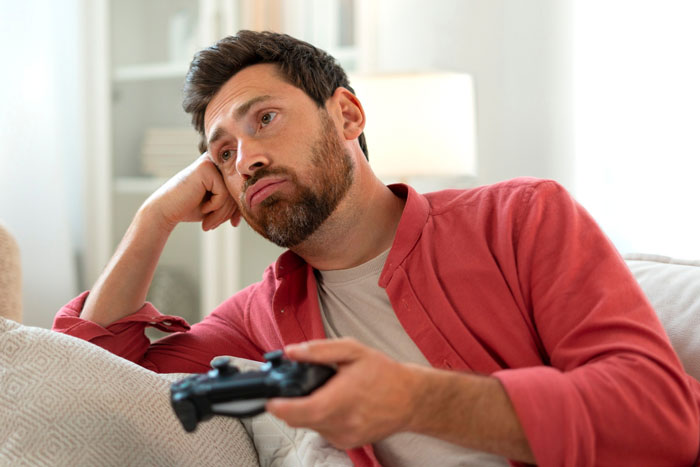 Manchild playing video games, looking bored and distracted while holding a game controller on a couch.