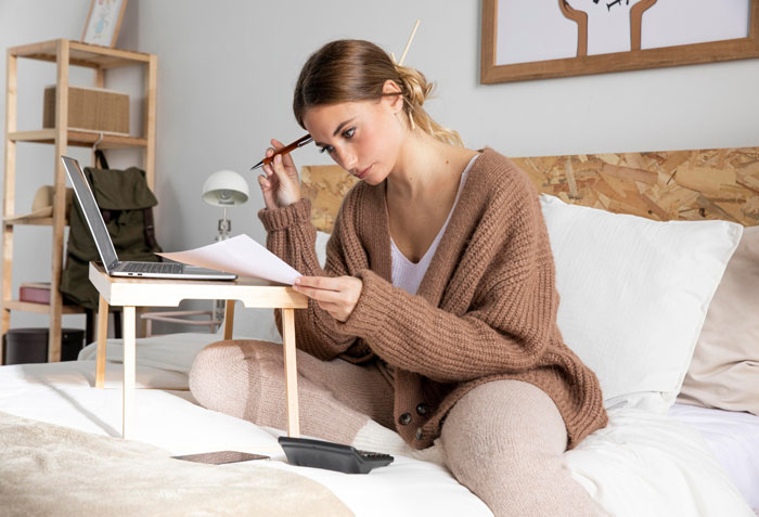 Woman in cozy clothes sitting on bed, reviewing expenses for birthday trip while using laptop and calculator nearby.