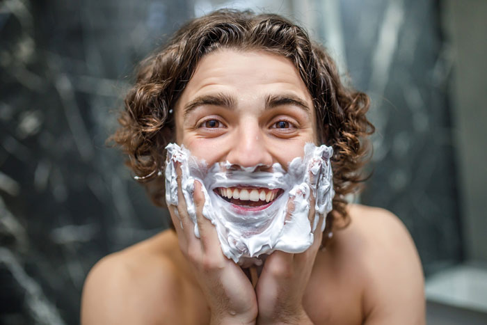 Young man with curly hair happily applying shaving cream on his face, using fancy skincare products in a bathroom. Young man with curly hair happily applying shaving cream on his face, using fancy skincare products in a bathroom.