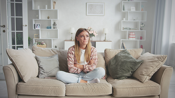 Young woman sitting on a couch with arms crossed, looking upset and contemplative in a bright living room.