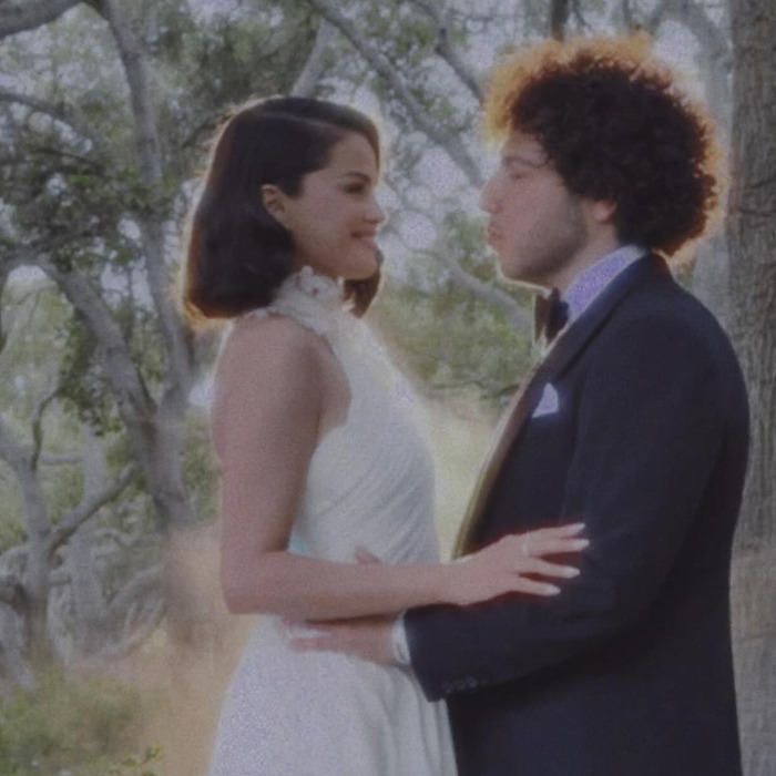 Selena Gomez in a white wedding dress embracing a man outdoors amid trees in a soft-focus intimate moment.