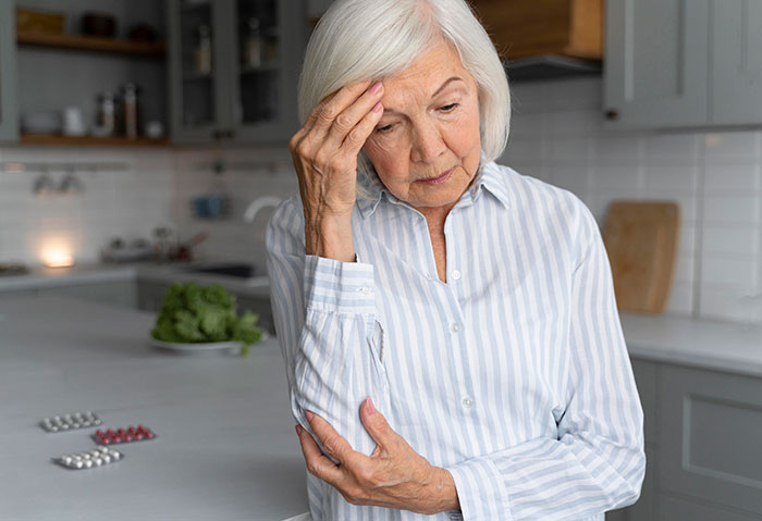 Older woman in striped shirt looking worried and stressed while standing in a kitchen near medication pills.