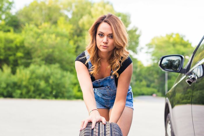 Young woman in denim overalls changing a car tire outdoors, illustrating drunk fumbles and bad decisions theme.
