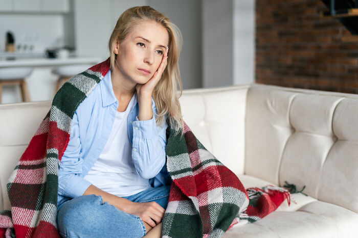 A woman wrapped in a blanket sitting on a couch, appearing thoughtful and affected by medical neglect.
