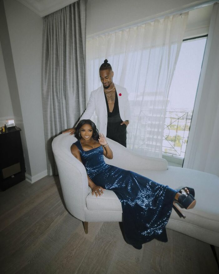 Simone Biles in a sparkling blue gown seated on a chaise lounge, with a man standing behind her in a white jacket indoors.