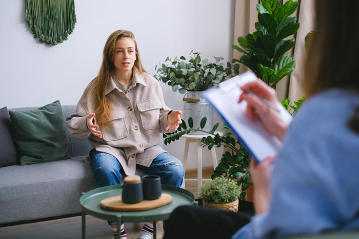 Young woman discussing family issues with therapist during session in a bright, plant-filled room focusing on ex-con mom struggles. Young woman discussing family issues with therapist during session in a bright, plant-filled room focusing on ex-con mom struggles.