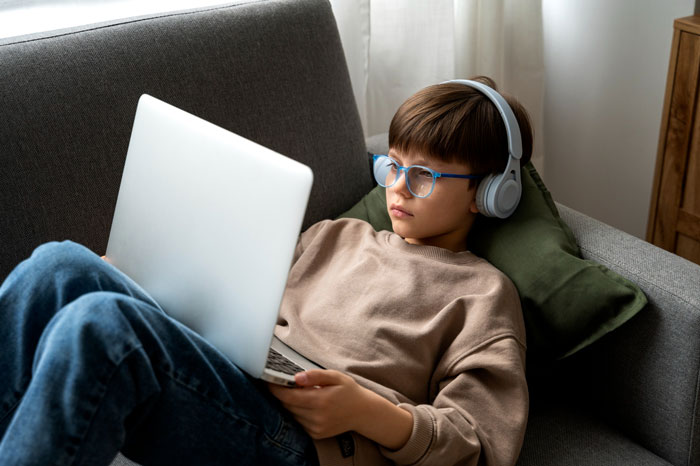 Young boy wearing headphones and glasses using a laptop on a couch, reflecting kids and ex-con mom family struggles. Young boy wearing headphones and glasses using a laptop on a couch, reflecting kids and ex-con mom family struggles.