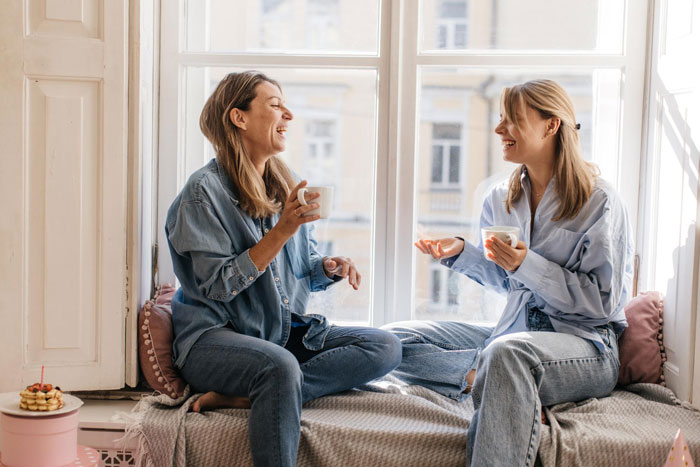 Two women talking and laughing while drinking coffee by a window, reflecting on family and ex-con mom challenges. Two women talking and laughing while drinking coffee by a window, reflecting on family and ex-con mom challenges.