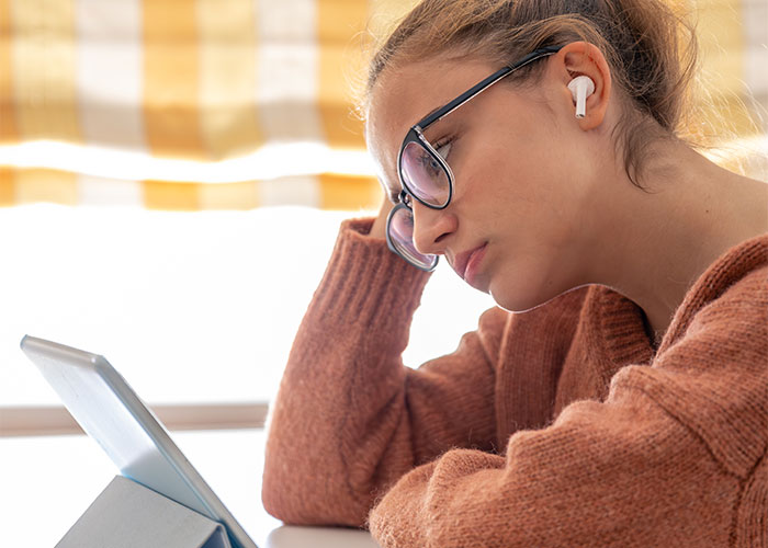 Young woman wearing glasses and earbuds, looking thoughtfully at a tablet, reflecting a golden child reality check.
