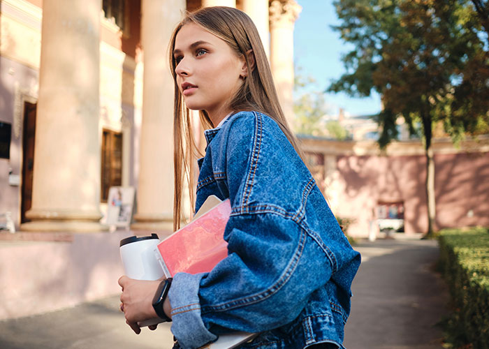 Young woman holding books outdoors near college campus, reflecting on golden child reality check after sister's success.