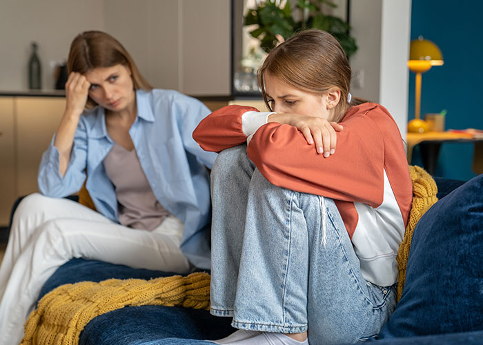 Two young women at home, one sitting curled up looking upset while the other watches with concern, capturing a reality check moment.