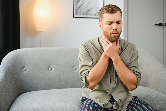Man sitting on a couch holding his throat in pain, illustrating unexpected hospital visits requiring surgery.