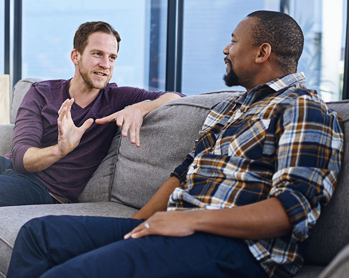 Two men sitting on a couch engaged in conversation demonstrating social cues that confuse people in communication.