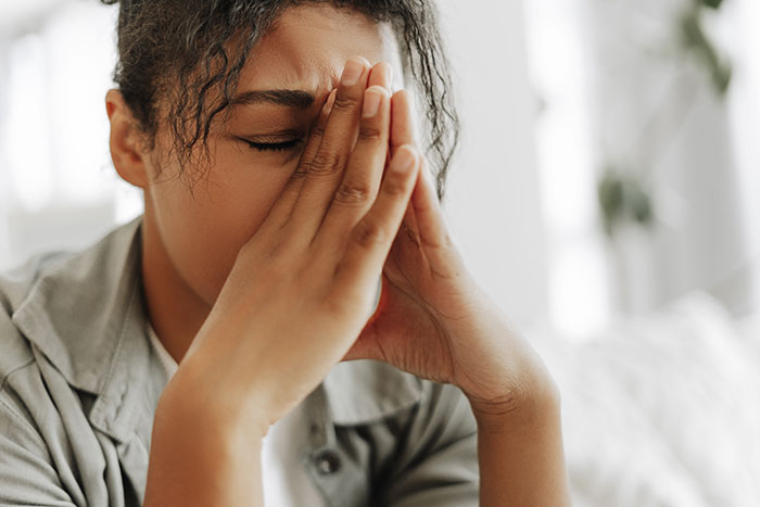 Young woman with eyes closed and hands pressed to face, showing confusion over social cues in a bright room.