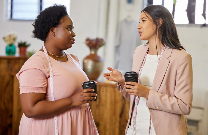 Two women holding coffee cups and discussing social cues that confuse them in a casual indoor setting.